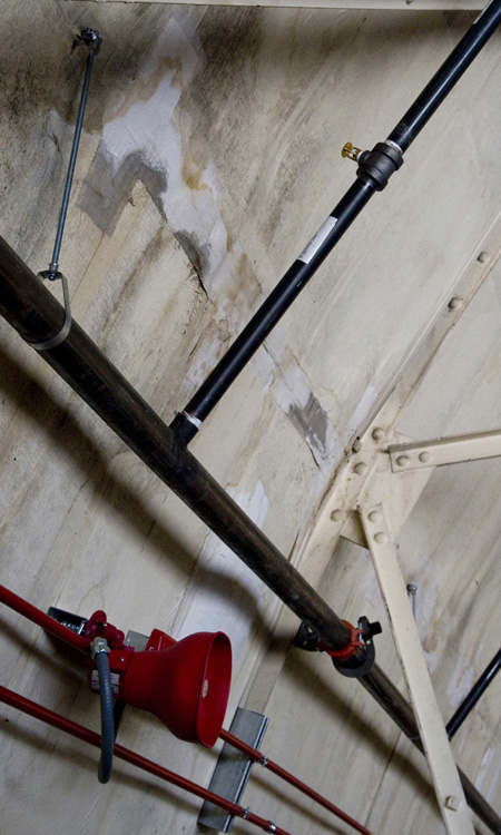 The newly installed foam fire suppression system on the wall of Hangar No. 1. Foam will spray from the nozzle at upper right; warning shrieks will sound from the loudspeaker at lower left.